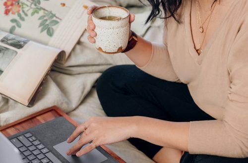 woman in beige long sleeve shirt and black pants sitting on bed using a Microsoft Surface Laptop 3