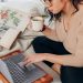 woman in beige long sleeve shirt and black pants sitting on bed using a Microsoft Surface Laptop 3